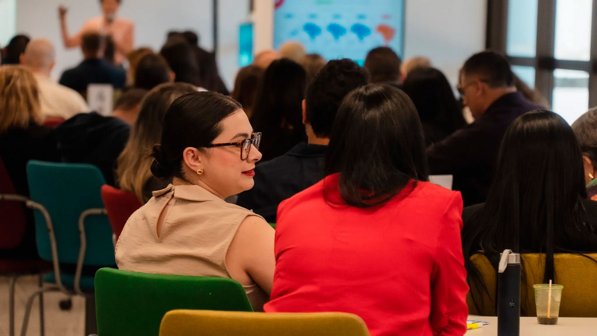 Um auditório com várias pessoas sentadas e olhando para a frente para uma tela ao fundo e desfocada. As pessoas são diversas em gênero e aparentam ser adultas. A iluminação é suave e o ambiente é de aprendizado.
