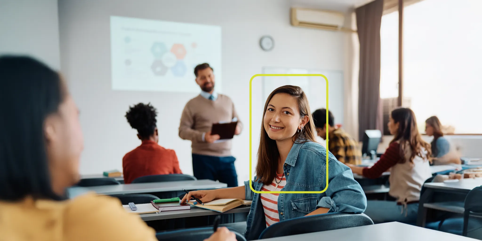 Uma jovem sorridente com cabelo castanho comprido e um casaco jeans está em primeiro plano à esquerda, olhando para a câmera. Ao fundo, desfocado, há outras pessoas e mesas em um ambiente de sala de aula ou escritório.