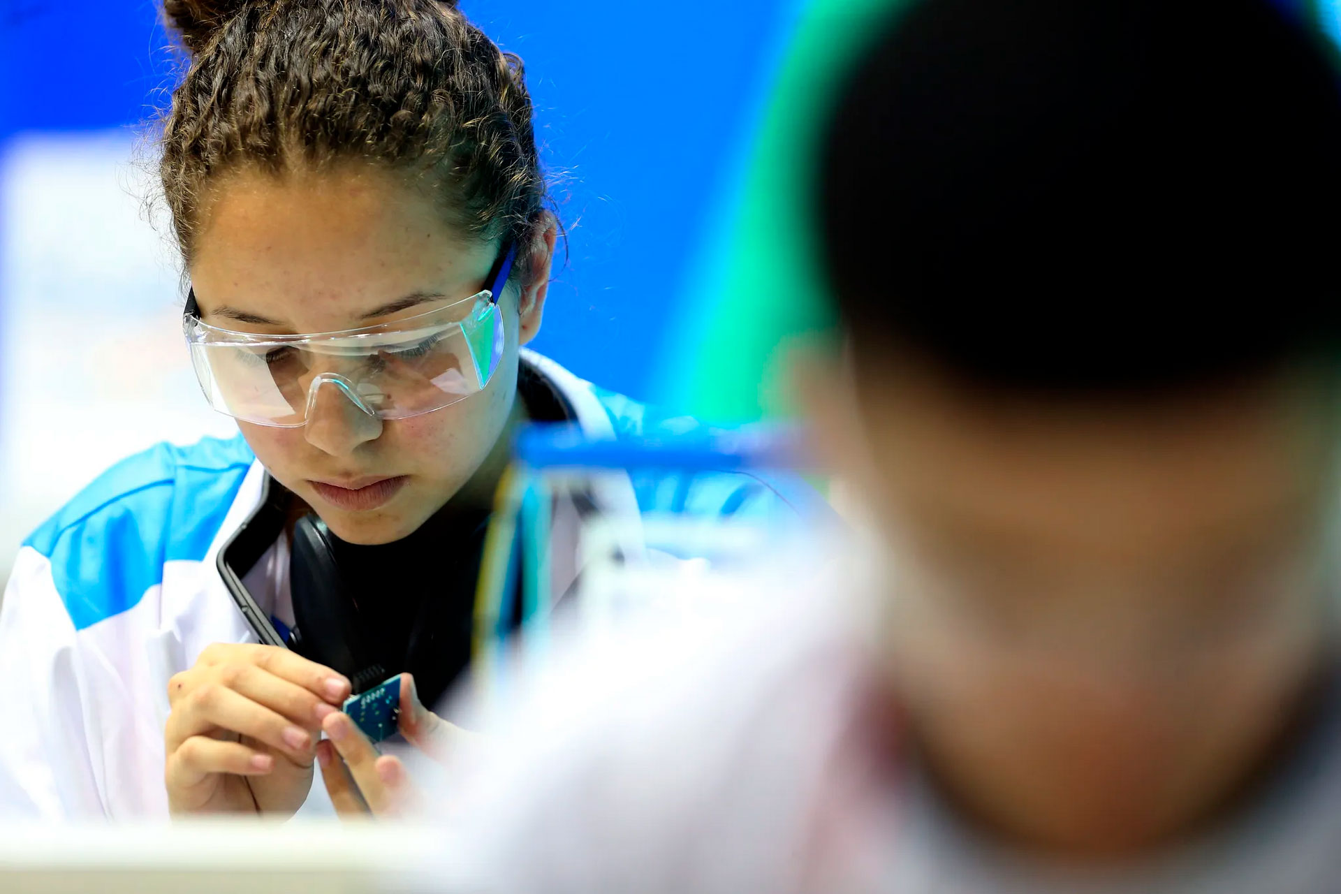 Uma jovem com óculos de proteção transparentes e cabelo preso está concentrada e trabalhando em um projeto. Ela veste um jaleco branco sobre uma camisa azul e branca. Parte da cabeça e ombro de outra pessoa são visíveis no primeiro plano, à direita. O fundo é desfocado em tons de azul.