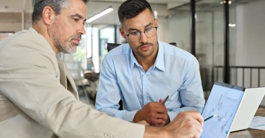 Duas pessoas em um ambiente de escritório estão próximas a um monitor grande que exibe gráficos e dados analíticos. O homem, à direita, gesticula em direção à tela. Ambos estão de pé e parecem engajados na discussão.