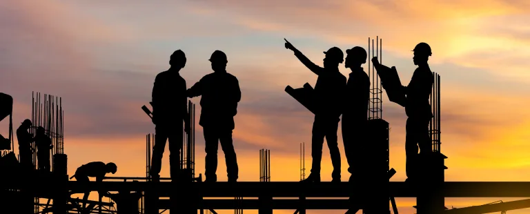 Silhueta de trabalhadores da construção civil sobre uma laje de concreto em um canteiro de obras ao amanhecer ou entardecer. Cinco trabalhadores estão em pé e um está abaixado, manuseando materiais ou ferramentas. Os trabalhadores em pé estão interagindo e um deles parece estar apontando para algo no distante. Estruturas de aço verticais indicam uma obra em progresso.