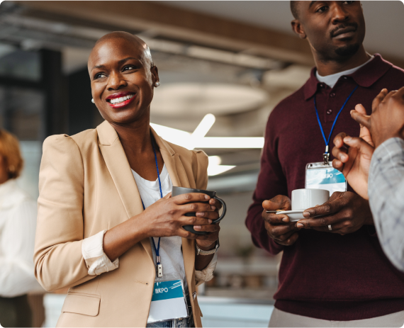 Três profissionais discutem animadamente enquanto tomam café em um ambiente de trabalho moderno. Uma mulher segura uma xícara e sorri.