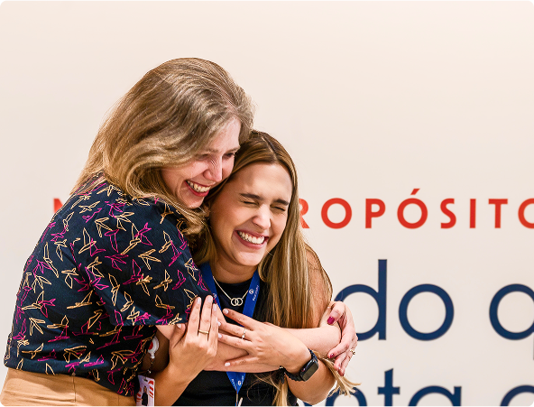 Duas jovens mulheres estão abraçadas e sorrindo calorosamente. A mulher à esquerda tem cabelos loiros ondulados e veste uma blusa estampada colorida. A mulher à direita tem cabelos castanhos longos e veste uma camiseta preta com um cordão azul no pescoço.