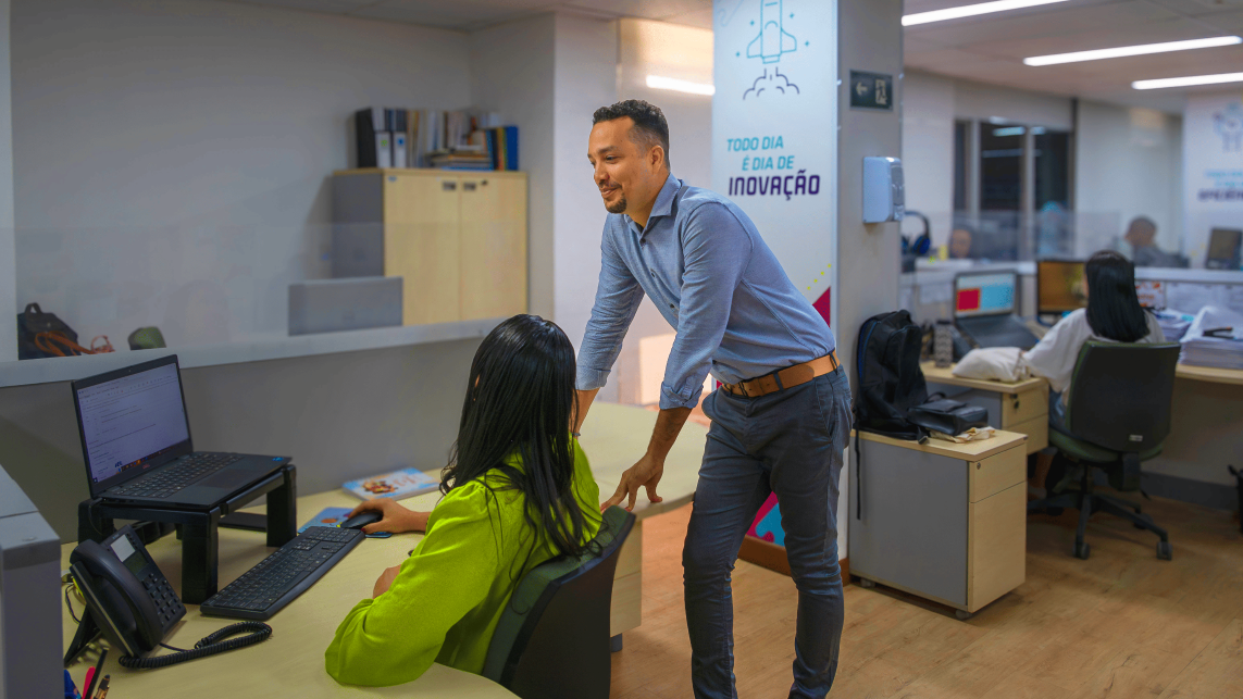 Em um escritório moderno com paredes brancas rabiscadas com diagramas e ideias em português, um homem com pele morena clara e camisa azul está em pé, gesticulando e falando com uma mulher com pele morena escura e blusa verde, que está sentada a uma mesa com um laptop.