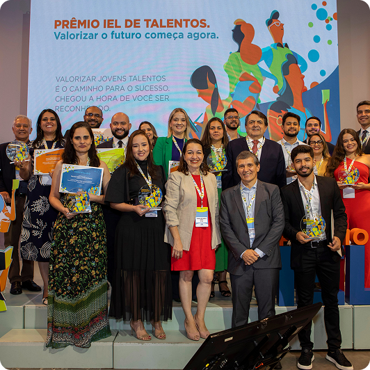 Um grupo diversificado de pessoas está posando para uma foto em um evento de premiação. Eles estão segurando troféus e sorrindo. Ao fundo, um banner vertical com texto e um logotipo ("PRÊMIO IEL DE TALENTOS") é visível. As pessoas são de diferentes idades e etnias, e estão em uma celebração.