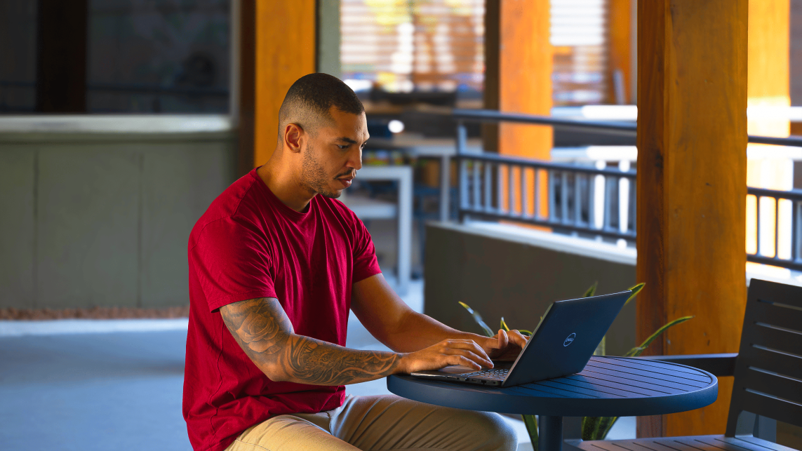 Um homem moreno, de camiseta vermelha e tatuagem no braço está sentado ao ar livre em uma mesa redonda, usando um laptop e uma caneta digital. O fundo mostra uma área externa com luz natural e elementos arquitetônicos.