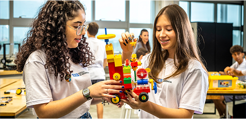 Duas pré-adolescentes em sala de aula seguram um projeto escolar, parecido com um carro.