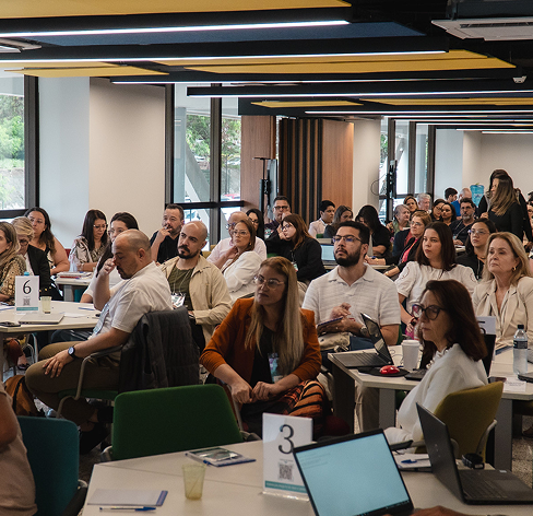 Grupo de professores sentados em mesas redondas em um auditório, assistindo a uma palestra. Eles estão participando de uma formação continuada do SESI.